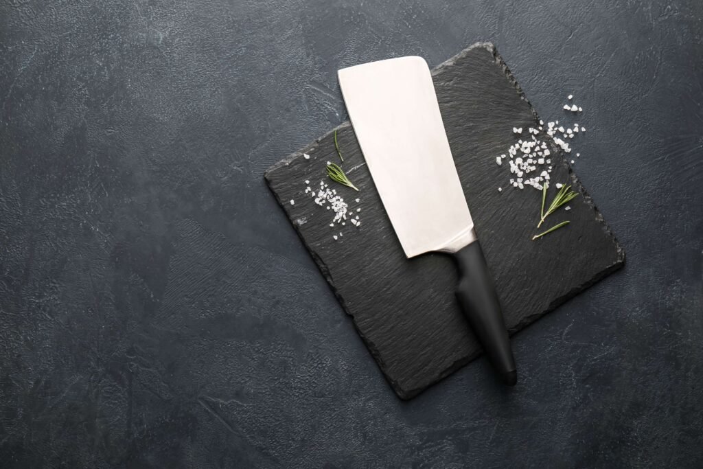 An image of a cleaver knife with a focus on the blade with salt grains and some green vegetables against a black background.